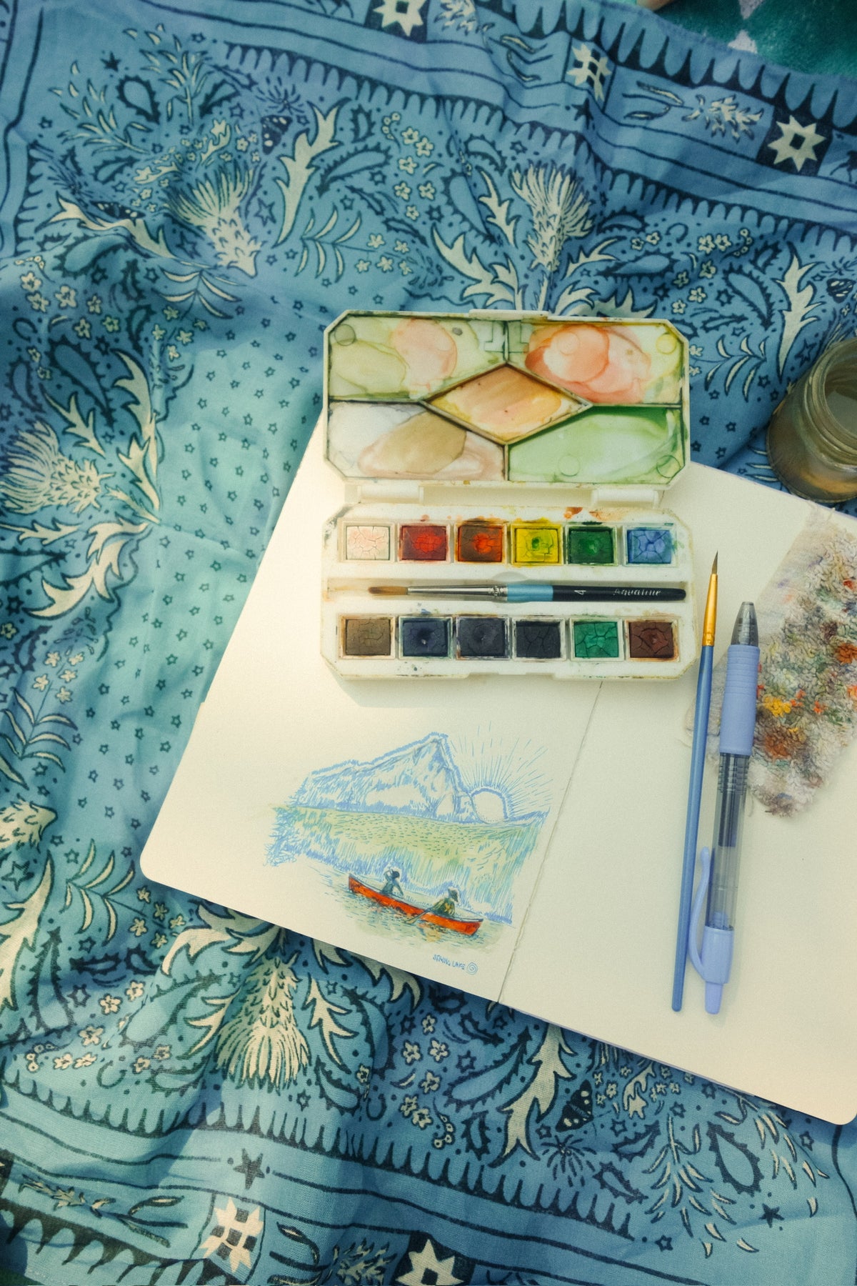 A watercolor paint palette with mixed paint colors, a small sketchbook with a landscape drawing, a blue pen, and a paint brush arranged on a blue patterned bandana background.