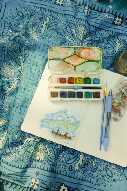 A watercolor paint palette with mixed paint colors, a small sketchbook with a landscape drawing, a blue pen, and a paint brush arranged on a blue patterned bandana background.