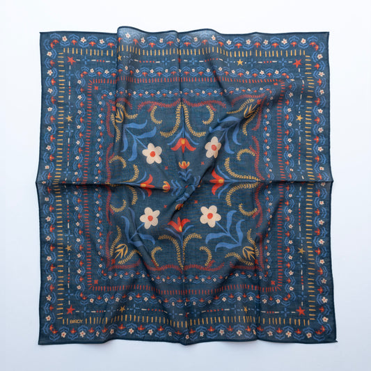 A square scarf with a dark navy blue base, featuring a detailed folk-art style border and central floral pattern in red, yellow, and white, laid flat on a white background.