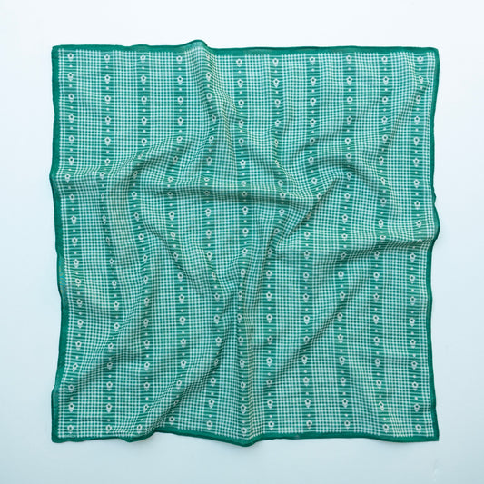 A square green and white gingham pocket square with vertical bands of small white floral motifs and a solid green border, laid flat on a light blue background.